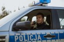 Zdjęcie w zbliżeniu policjanta w nowym umundurowaniu, który siedzi za kierownicą radiowozu oznakowanego i przy ustach trzyma mikrofon do mówienia przez stację z dyżurnym.
